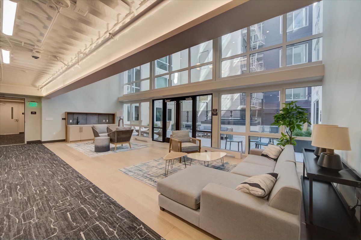 Lounge area with large windows, light wood floors, gray sectional sofa, armchairs, and side table with lamp.