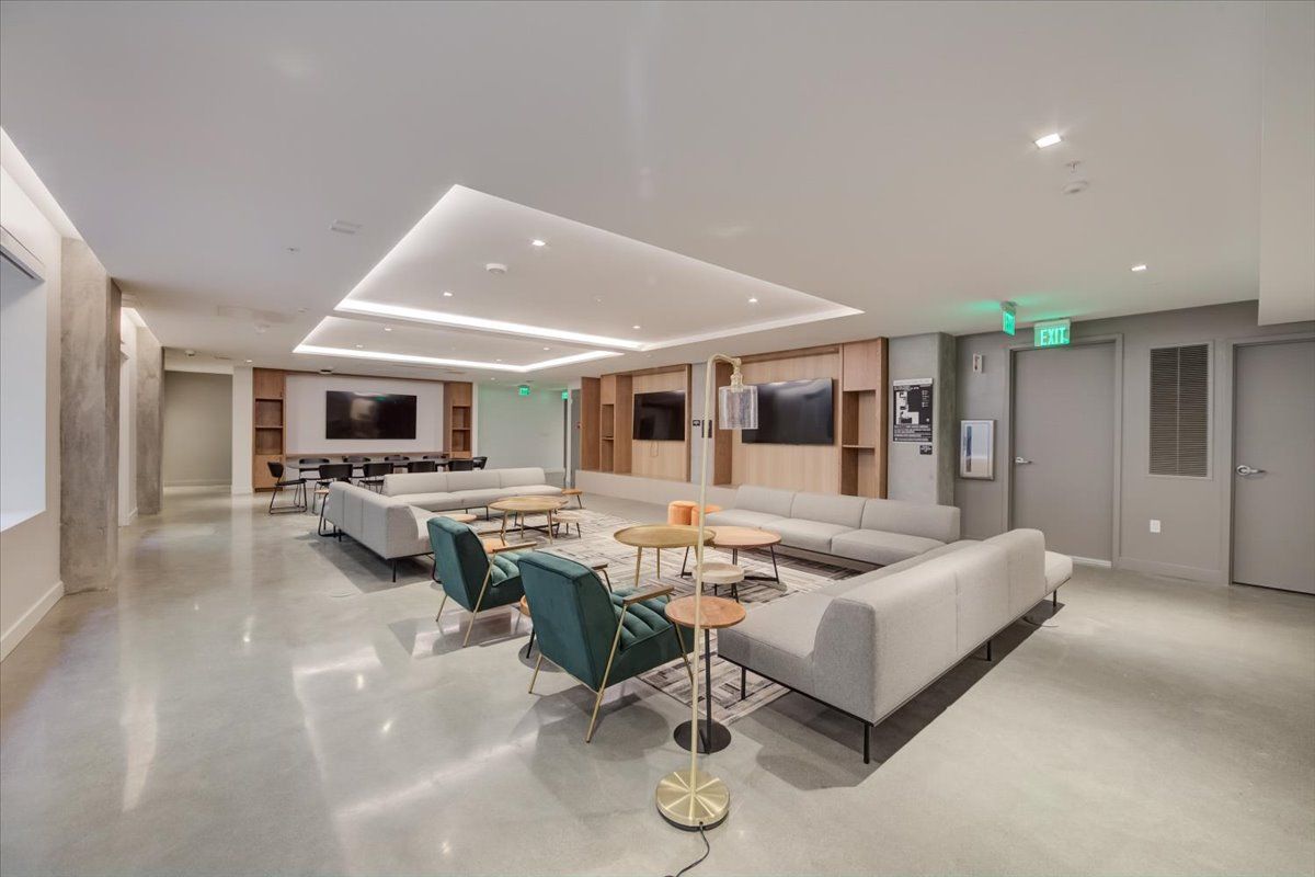 Lounge area with light gray couches, teal armchairs, and wood accent walls. Two TVs and small tables. Shiny concrete floor.