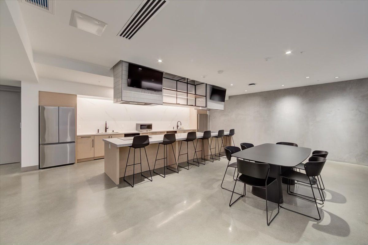 Modern break room with kitchen, bar seating, table with chairs, and two TVs. Gray, white, and wood tones.