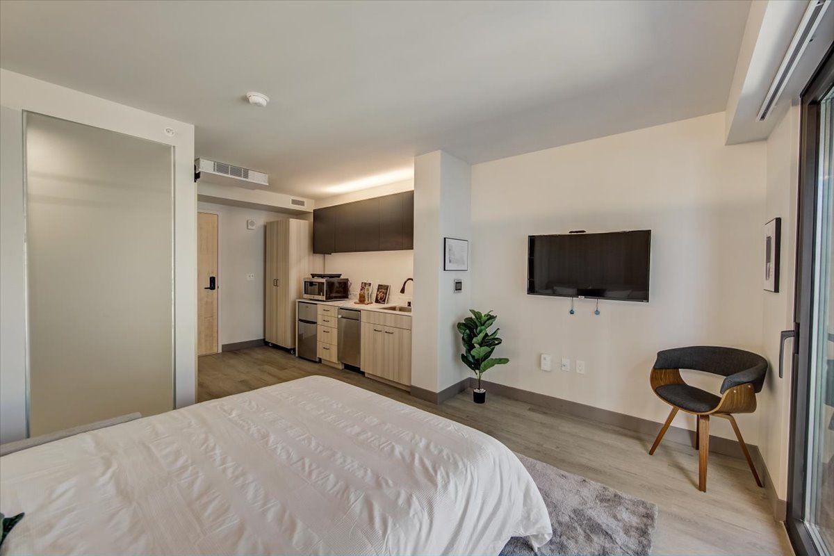 Bedroom interior with bed, TV, kitchen area, and a chair. Light color scheme.