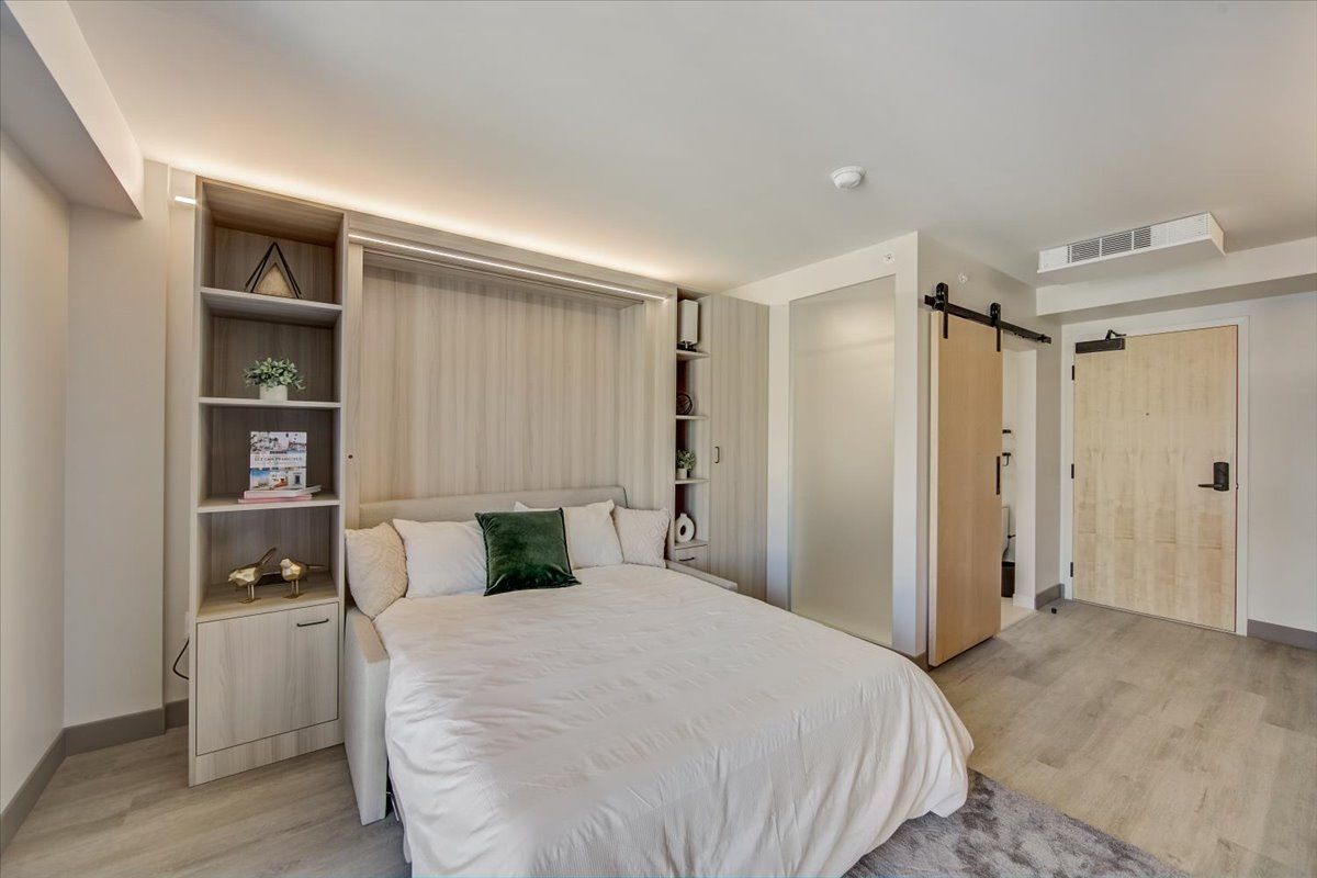 A small bedroom with a bed, shelving, and a sliding door. Light wood and white accents.