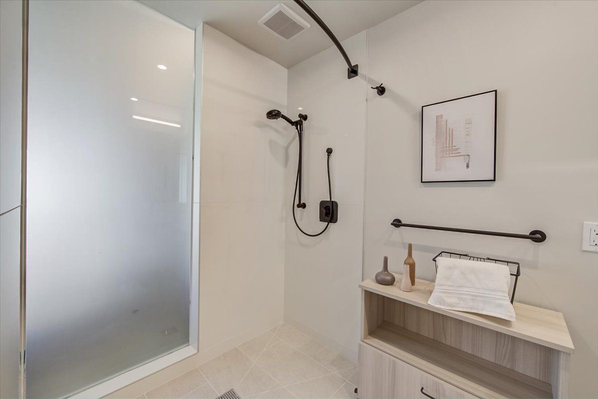 Modern bathroom with walk-in shower. Frosted glass panel, black shower fixtures, light wood storage, and decorative wall art.