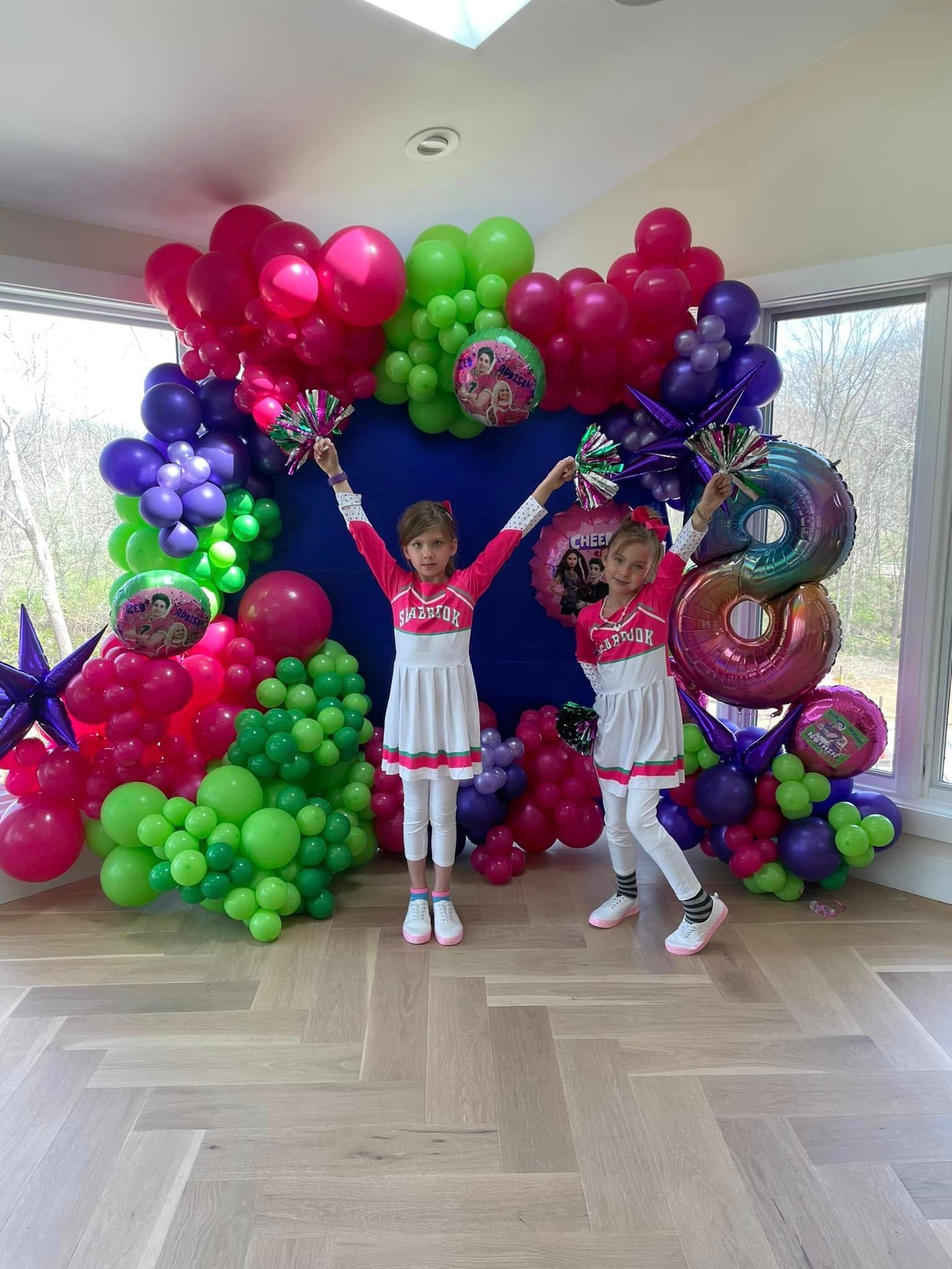 Two little girls are standing in front of a wall of balloons.