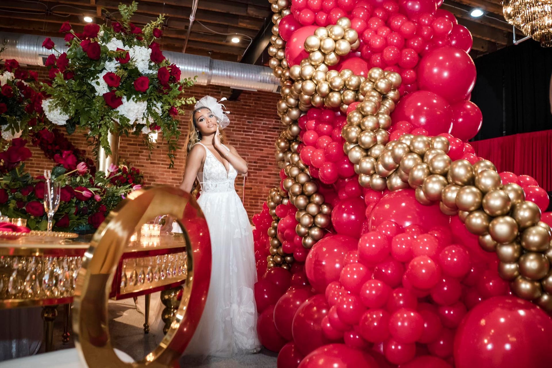A bride in a white dress is standing in front of a wall of red and gold balloons.