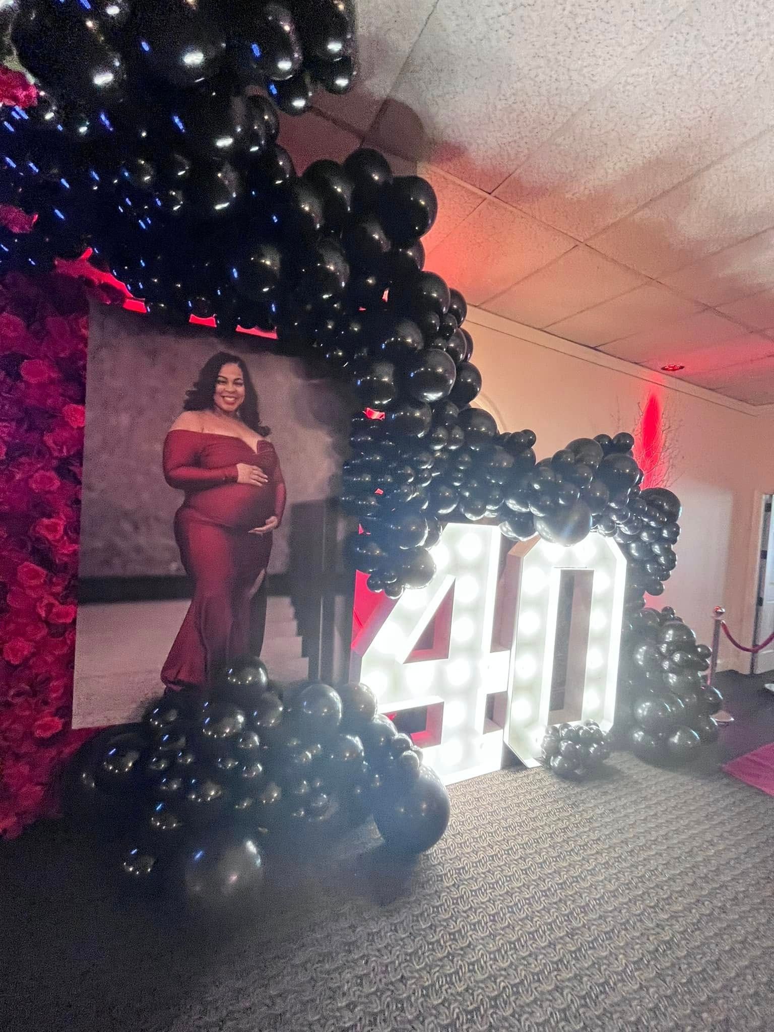 A pregnant woman is standing in front of a 40 sign surrounded by black balloons.