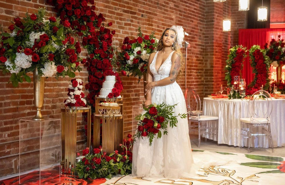 A bride in a white dress is standing in front of a brick wall holding a bouquet of red roses.