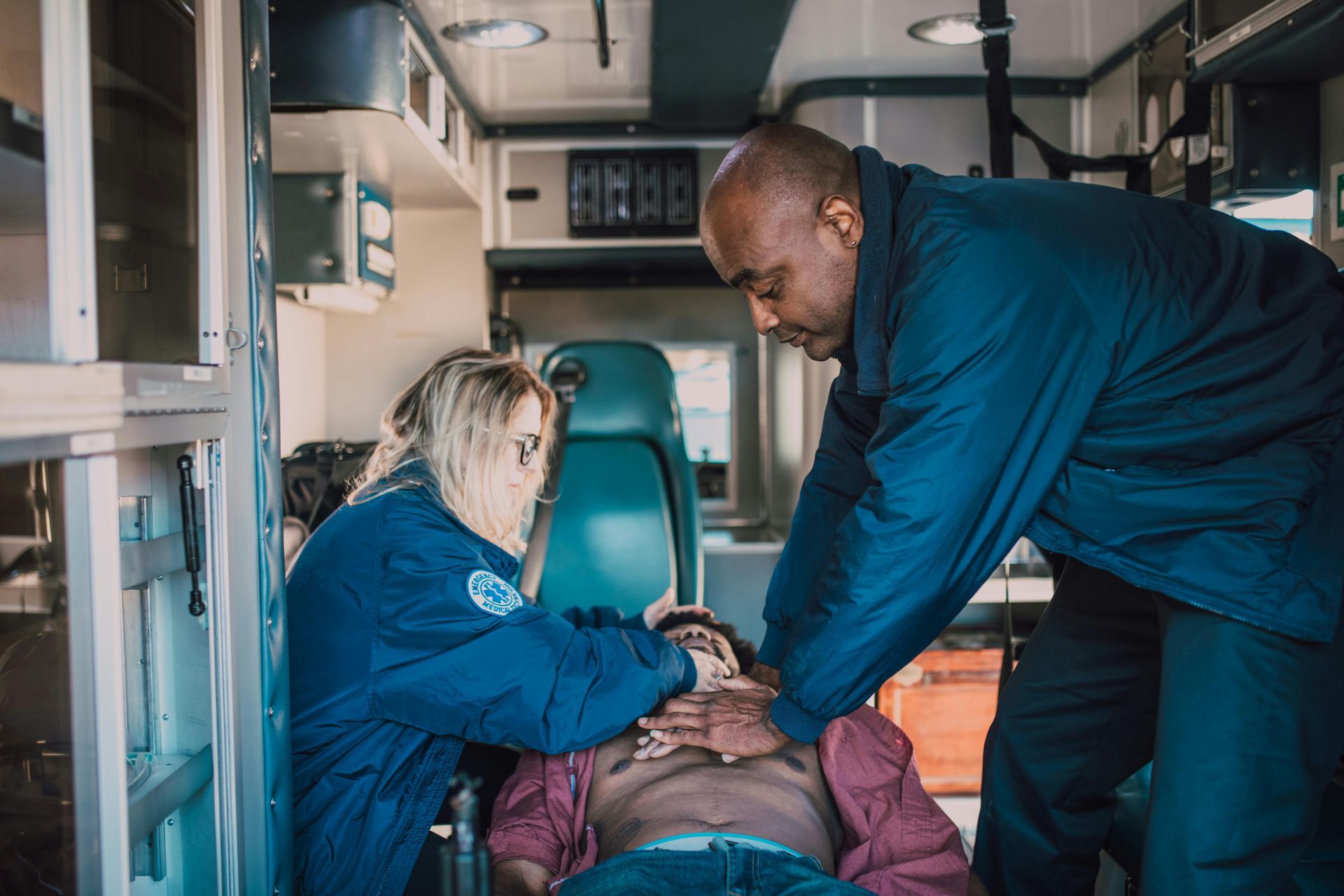 Woman in blue scrubs performs CPR on a person lying on a mat in a classroom.