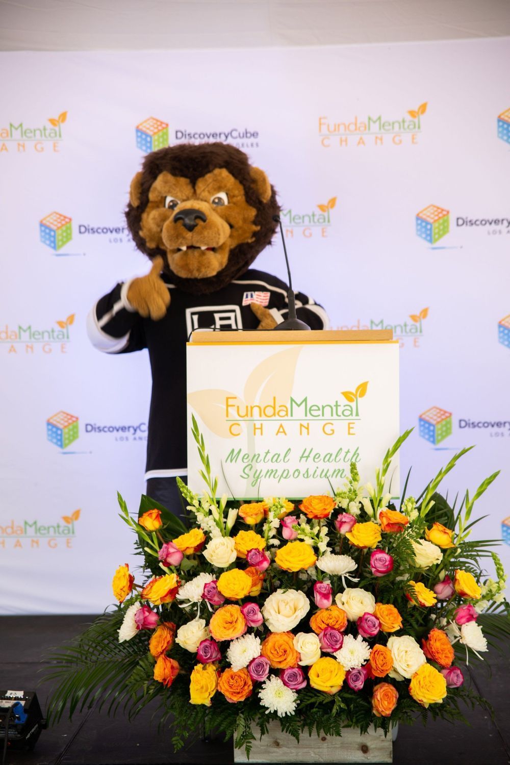 A lion mascot in LA Kings jersey stands at a podium with flowers, promoting Mental Health Symposium.