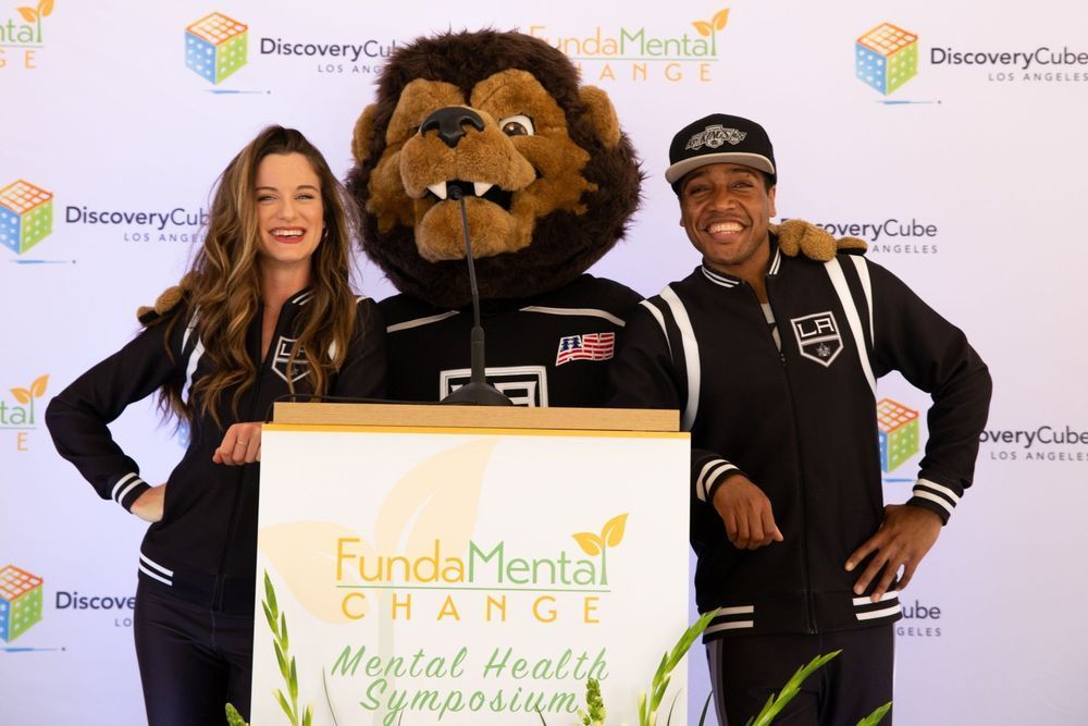 A woman, mascot, and man in black jackets stand at a podium for a mental health symposium.
