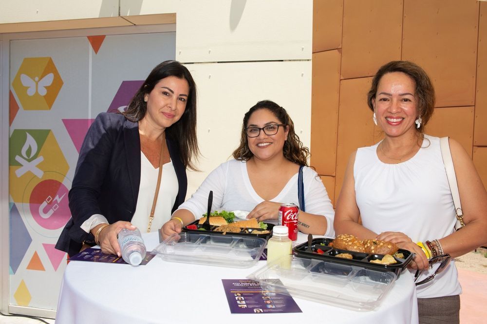 Three women at a table with food. Two are smiling. A butterfly design is on the wall.