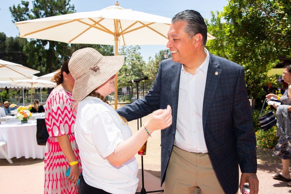 Man in blazer smiles, interacts with person in sun hat, other people and tables in background.