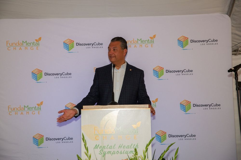 Man at podium speaking, gesturing with hands. Backdrop with logos and text,