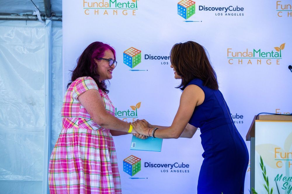 Two women shaking hands in front of a backdrop with logos. One wears pink plaid, the other blue.