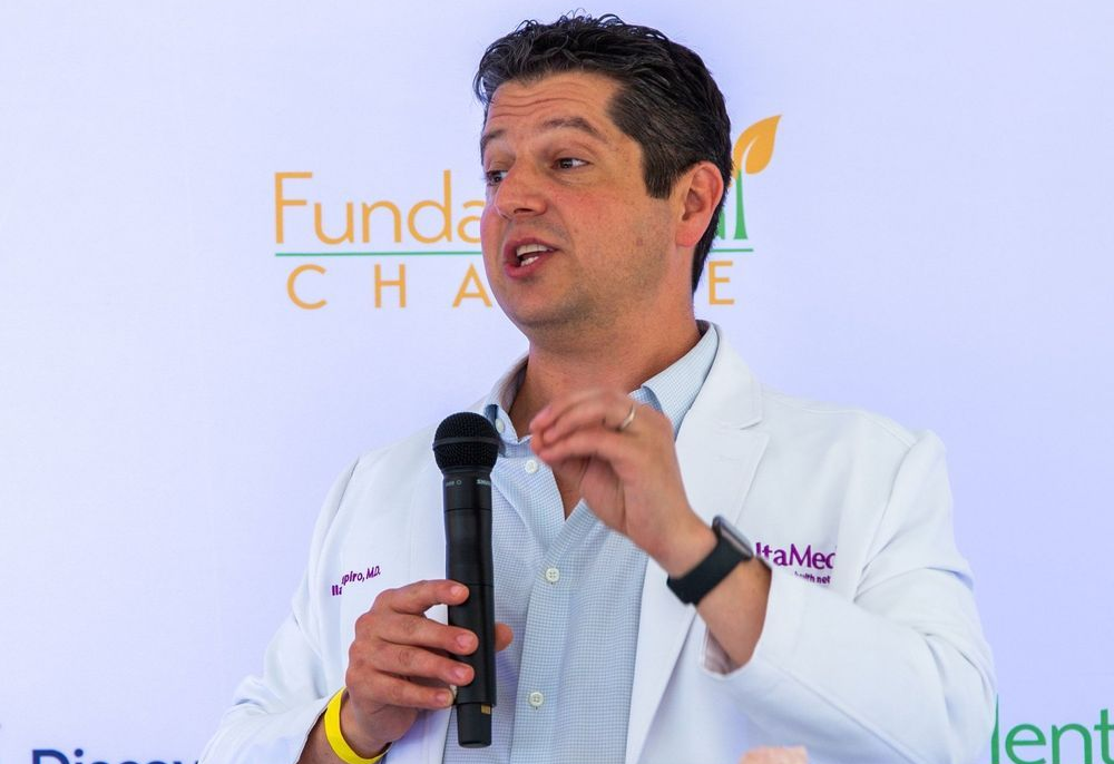 Man in white coat speaking into microphone; green and orange banner in background.