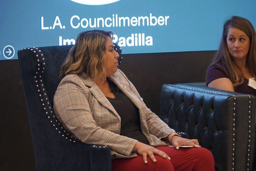 Woman in a blazer speaking, sitting on a black tufted chair next to another woman at a panel.