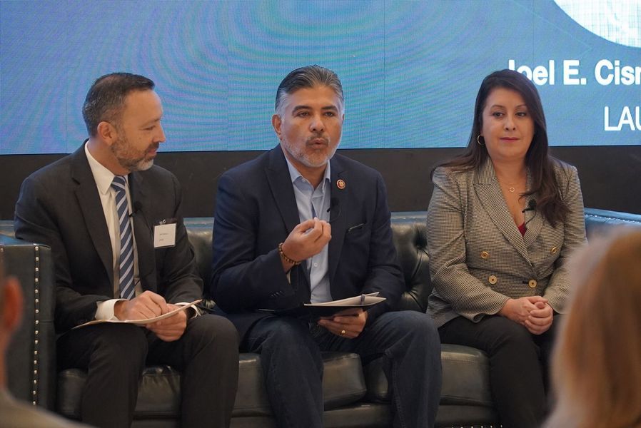 Panel discussion with three people on a black couch, speaking and taking notes, in front of a screen.
