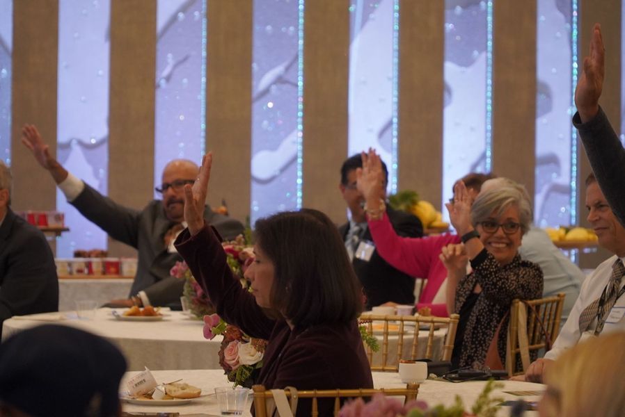 People at a table raise their hands, likely in a meeting or event, with an elegant background.