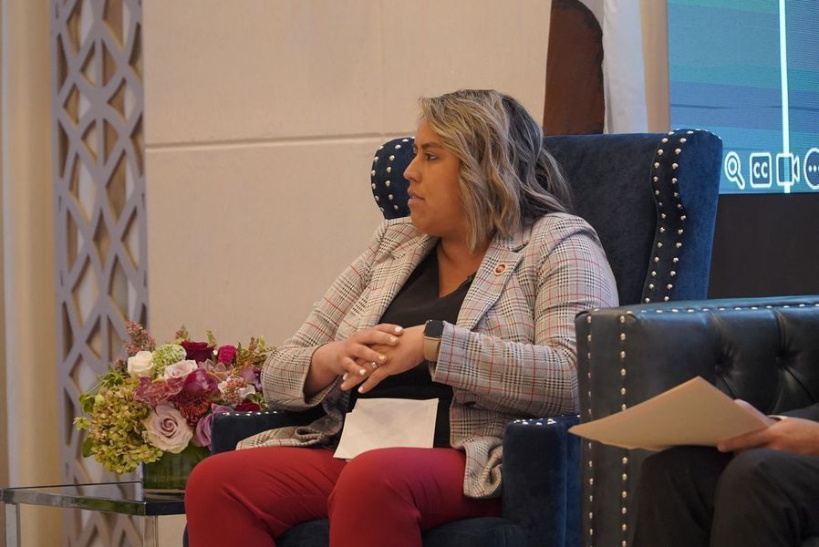 Woman in a blazer sits on a blue upholstered chair, facing left. She has her hands clasped.