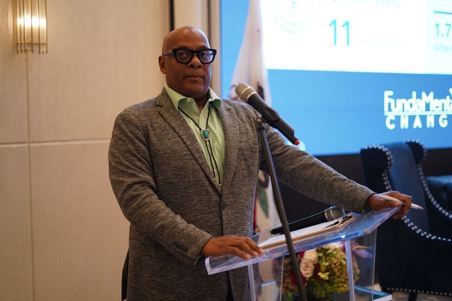 Man speaking at a podium, wearing a grey jacket and green shirt. A presentation is in the background.