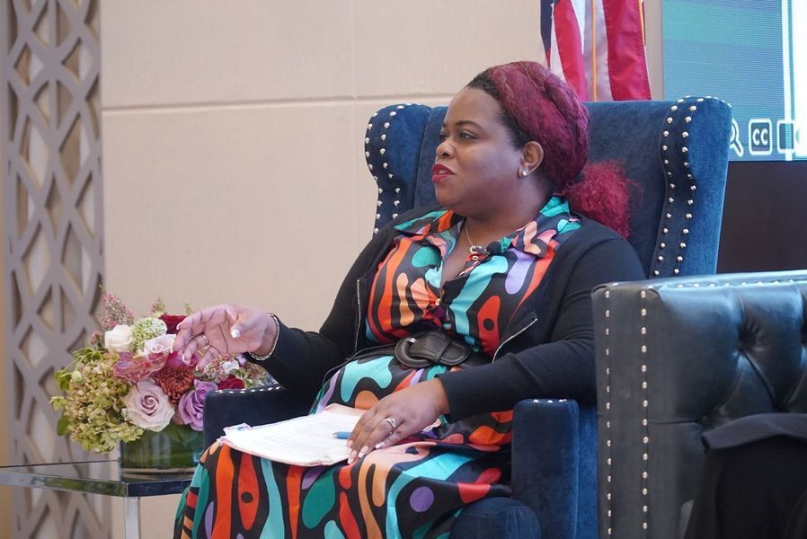 Woman in colorful dress and black cardigan speaks at event, seated in blue armchair.