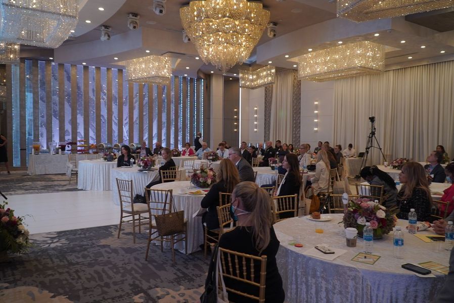 Large event in a ballroom with attendees at round tables, chandeliers, and a stage area.