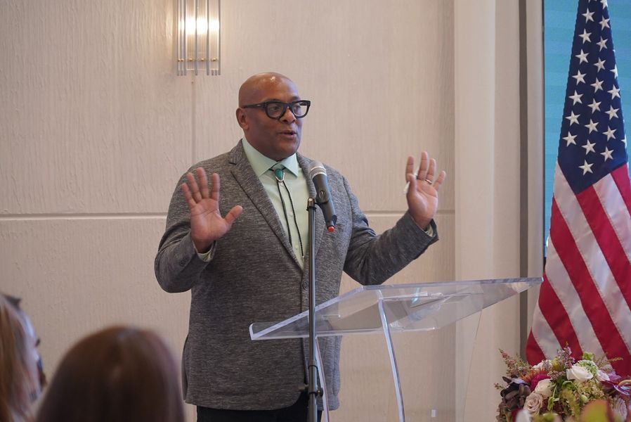Man speaking at a podium, gesturing with hands. American flag in background.