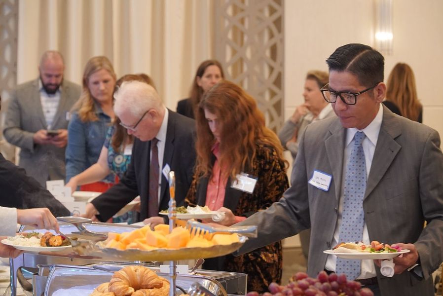 People at a buffet, selecting food. Indoors, neutral colors, some wearing name tags.