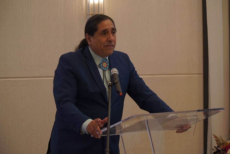 Man in a blue suit speaks at a podium with a microphone. He has long hair and a bolo tie.