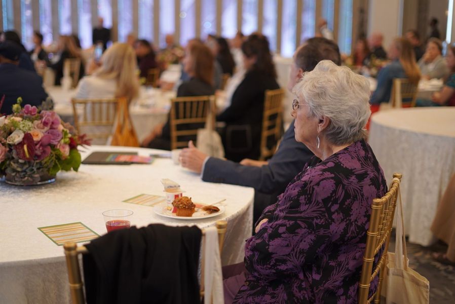 People seated at round tables in a well-lit room, observing an event; a woman in purple looks on.