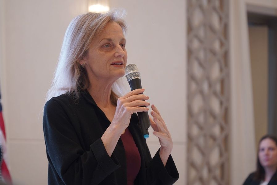 Woman speaking into a microphone, gesturing with her hands. Indoors, facing a crowd.