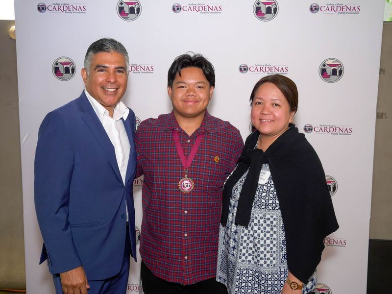 Three people stand in front of a backdrop with a logo. Man in blue blazer, center man with medal, woman in patterned dress.