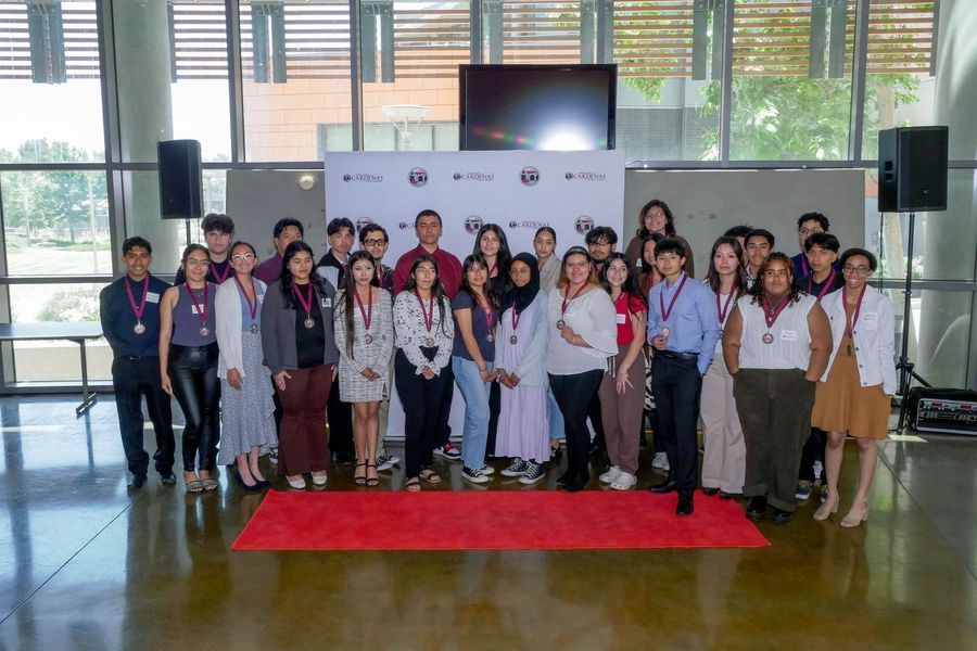 Group poses on red carpet at an event. People wear medals, stand in a bright, modern setting.