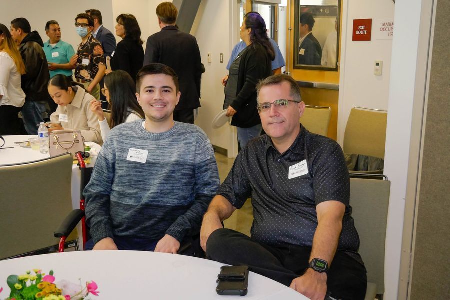 Two men seated at a round table, smiling, with a crowd in the background at an event.