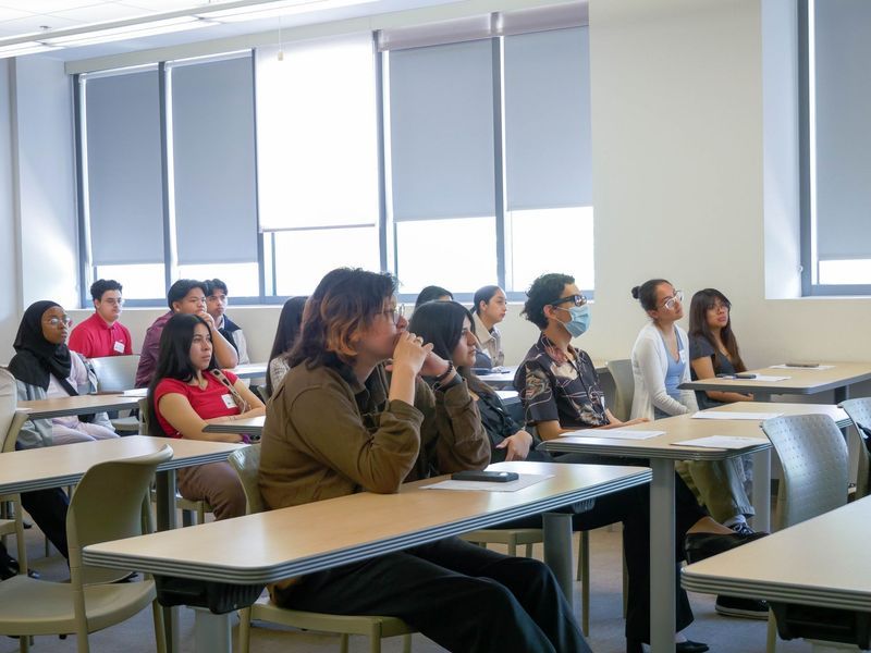 Classroom of students listening attentively. Desks and windows visible. Several students looking forward.