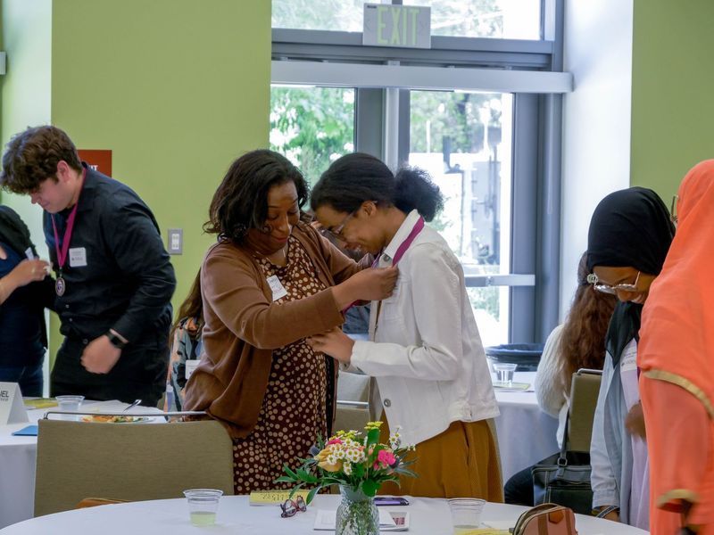 Woman pinning a badge on another person in a light-filled room with others at tables.