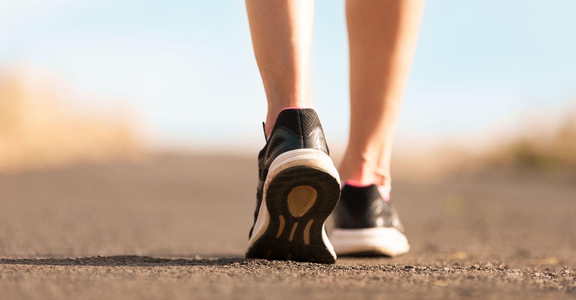 Person's feet in sneakers walking on a paved path outdoors; shallow focus on the shoes.
