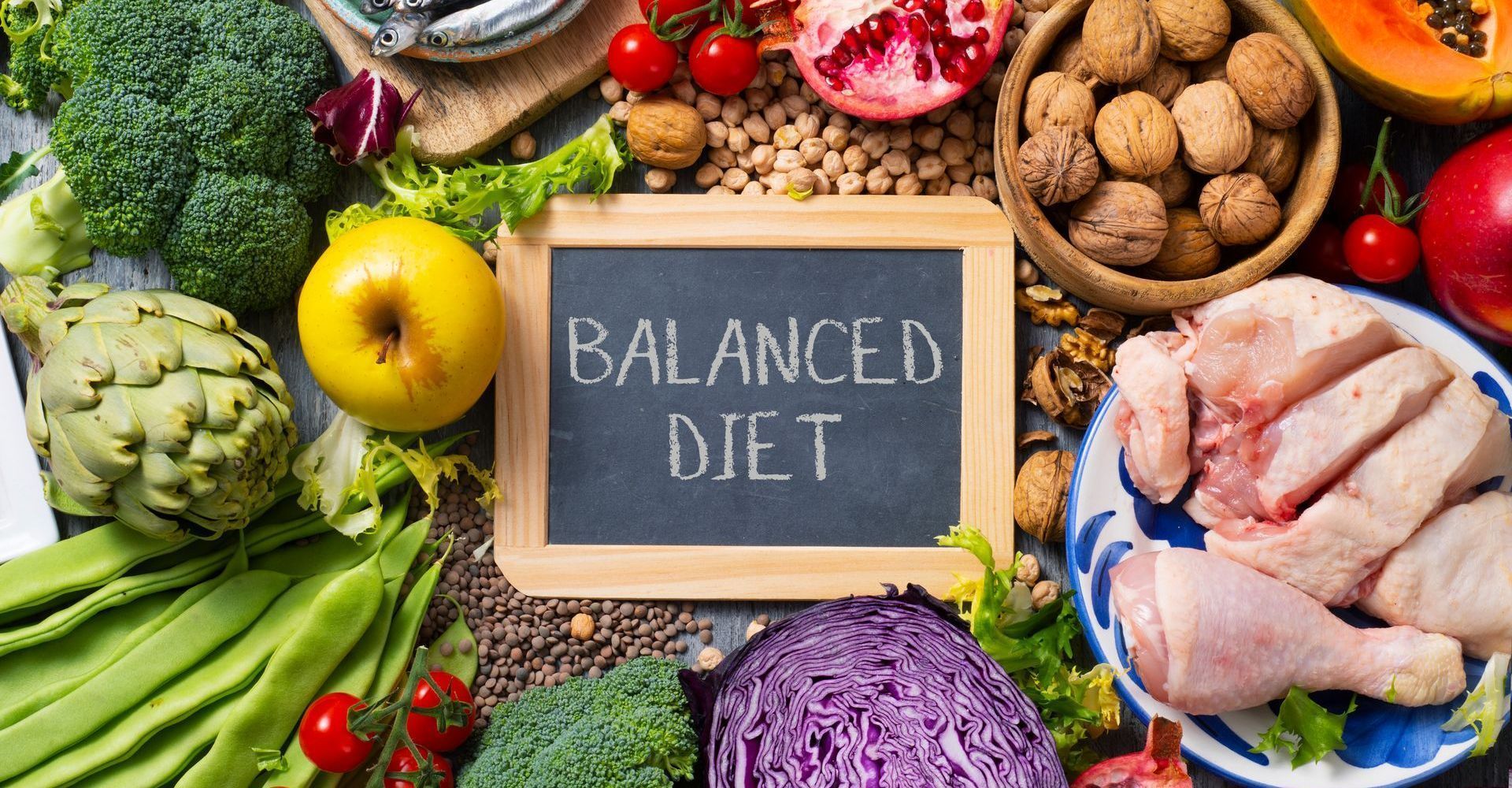 A variety of colorful fruits, vegetables, nuts, and chicken are arranged around a chalkboard that reads