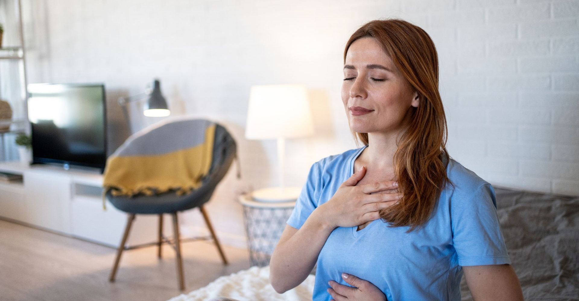 Woman with eyes closed, hands on chest and stomach, practicing deep breathing indoors.