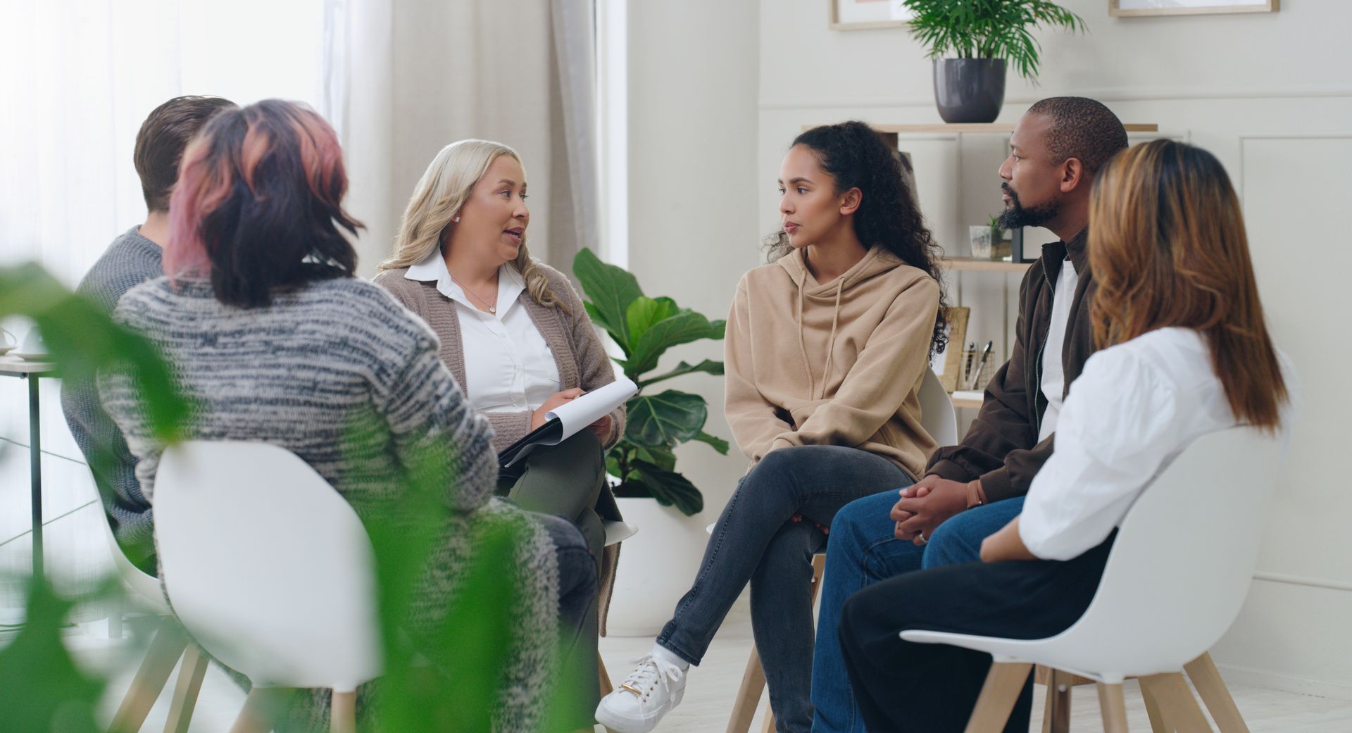 People sitting in a circle, engaged in a discussion; interior setting with plants.