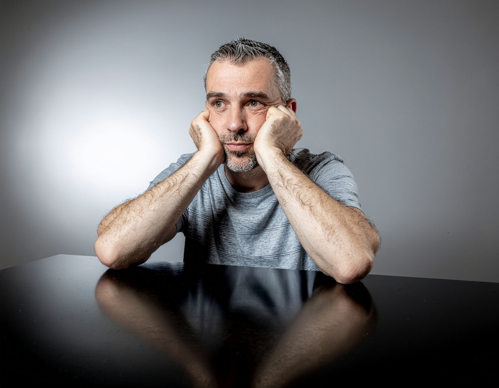 Man with graying hair rests chin on hands, looking despondent at a dark table.