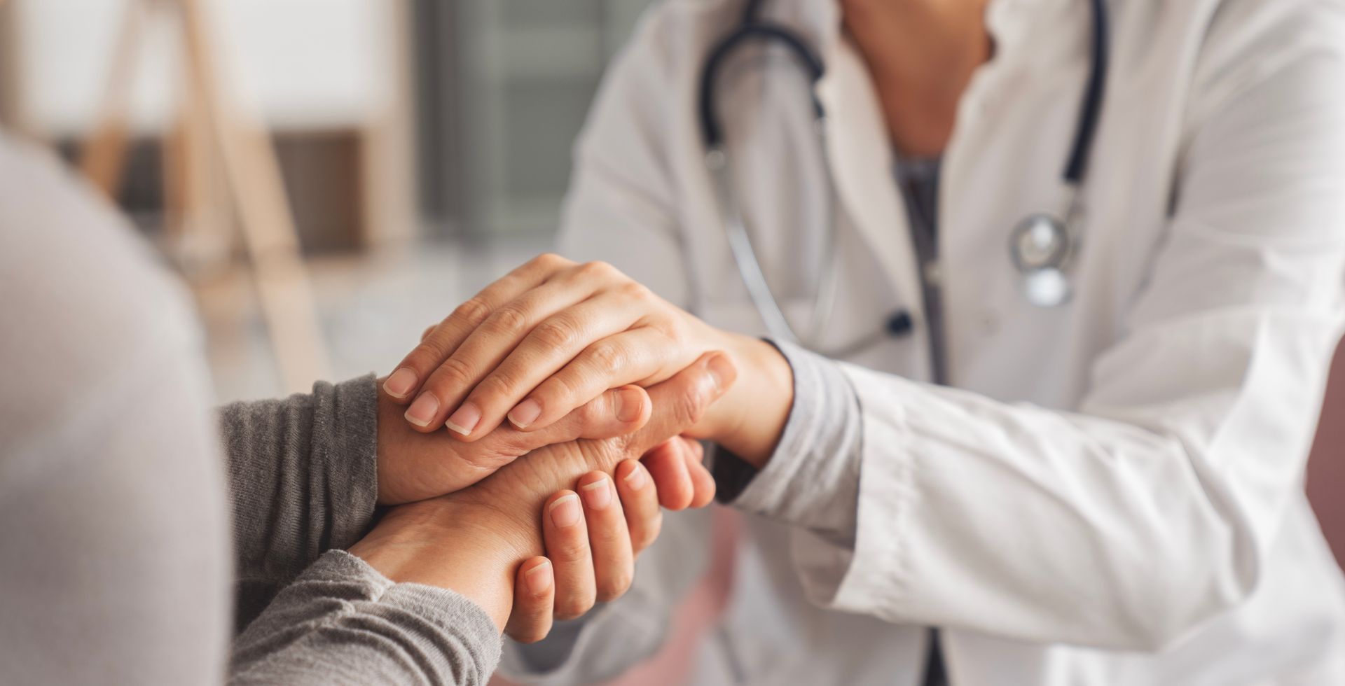 Doctor holding patient's hands, offering comfort. White coat, stethoscope. Indoor setting.