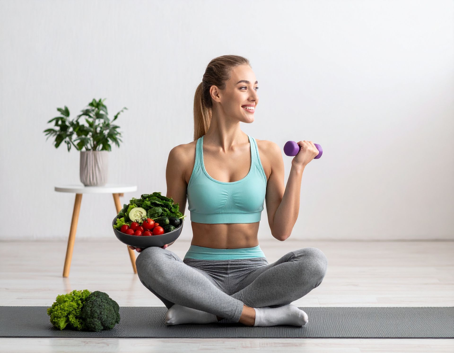 Woman in workout clothes with bowl of vegetables and dumbbell, smiling.