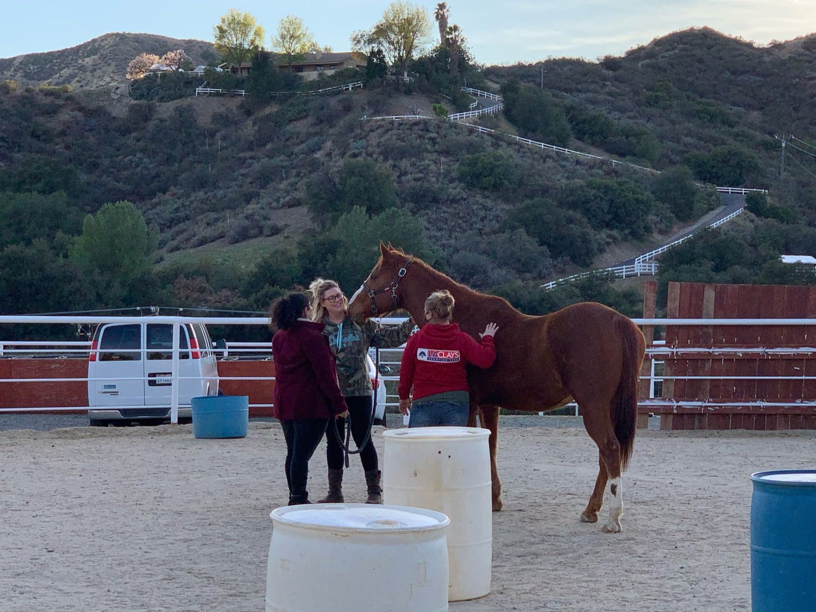Three people interacting with a chestnut horse in an outdoor arena, barrels nearby, hills in background.