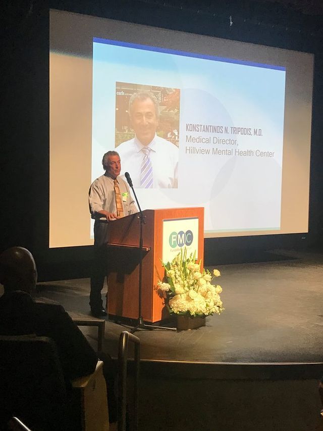 A man speaks at a podium with a screen behind him showing another man. White flowers decorate the podium.