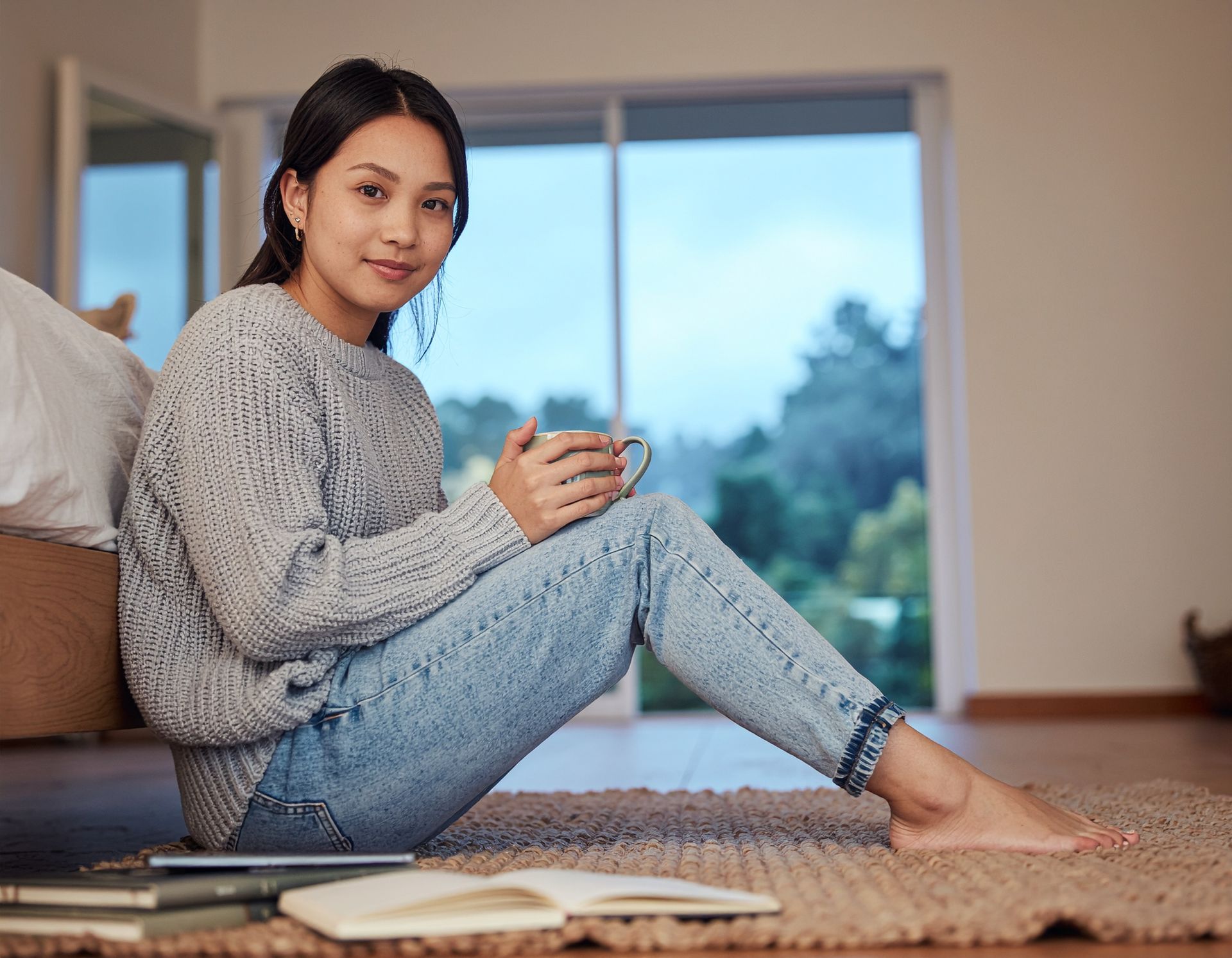 Person sitting on a rug with a mug, near an open book, in a cozy living room by a window.