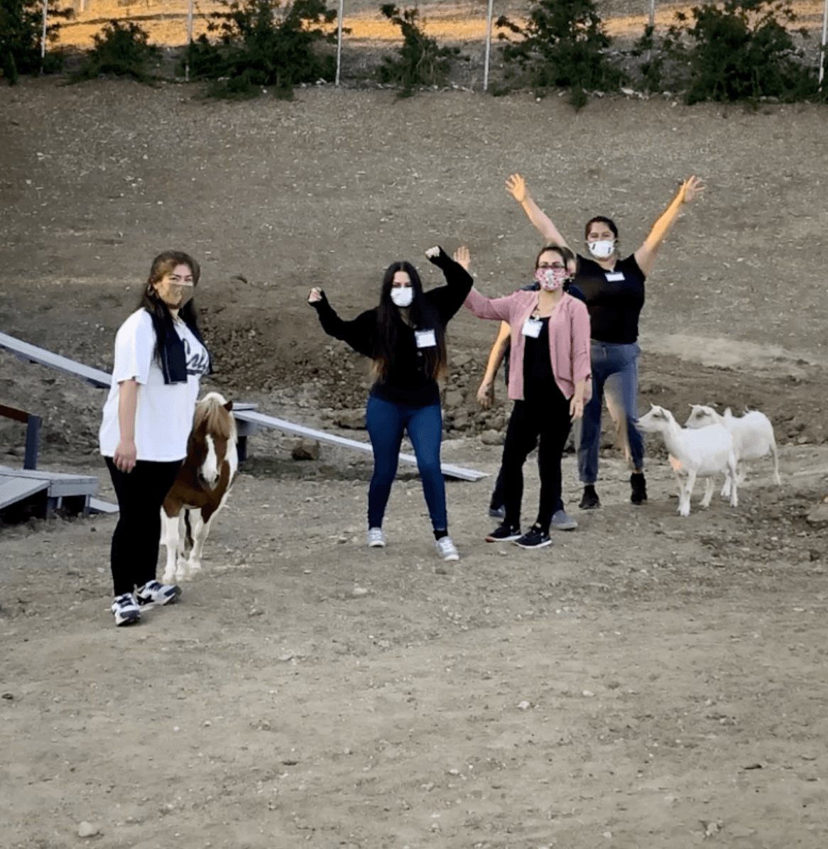 Four people and two goats posing outdoors. People are wearing masks and raising arms.