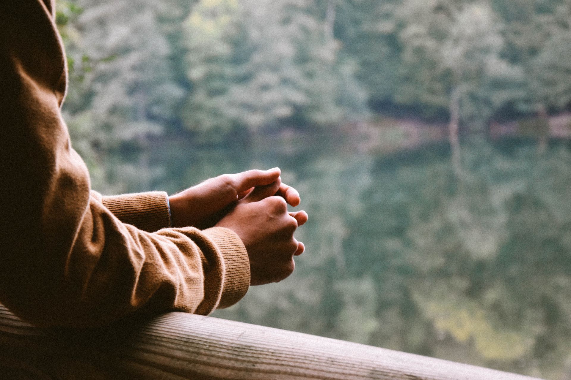 Person’s hands clasped on wooden railing, looking out at a tranquil lake with trees reflecting in the water.