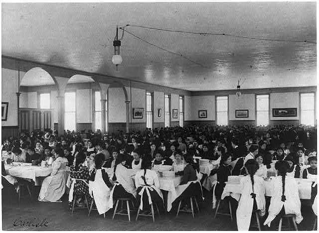 Large room with numerous people seated at tables, likely a dining hall. White tablecloths, overhead lights.