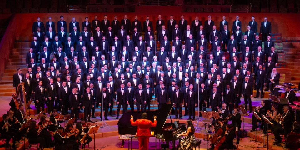 A large choir and orchestra perform on a stage; the conductor wears a bright orange suit.