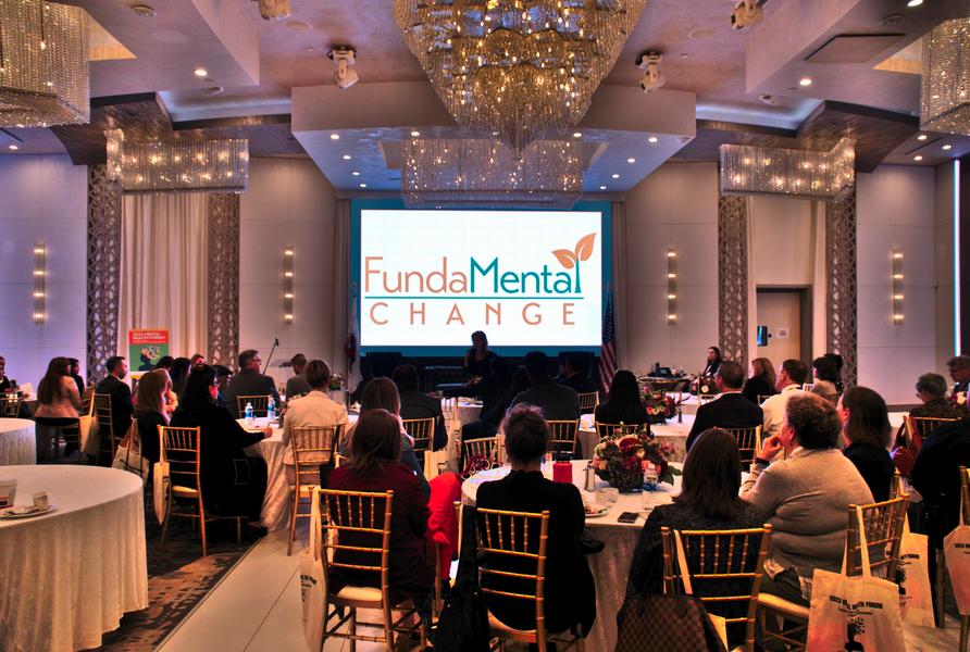 A conference room with attendees seated at tables, facing a screen displaying
